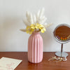 A pink ceramic bud vase holds fluffy white pampas grass and yellow flowers on a wooden surface.