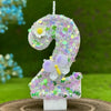 A large purple number cake candle decorated with small flowers and a butterfly on a green outdoor background.