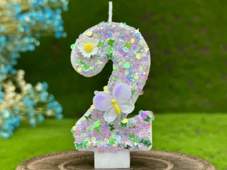 A large purple number cake candle decorated with small flowers and a butterfly on a green outdoor background.