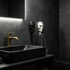 A black vessel sink and faucet are set against a dark gray tiled wall with a mounted skull hook and a towel.