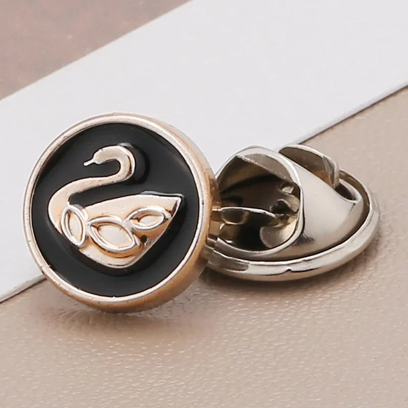 A pair of silver-toned no-sew brooch buttons featuring a black and gold design with a stylized swan motif.