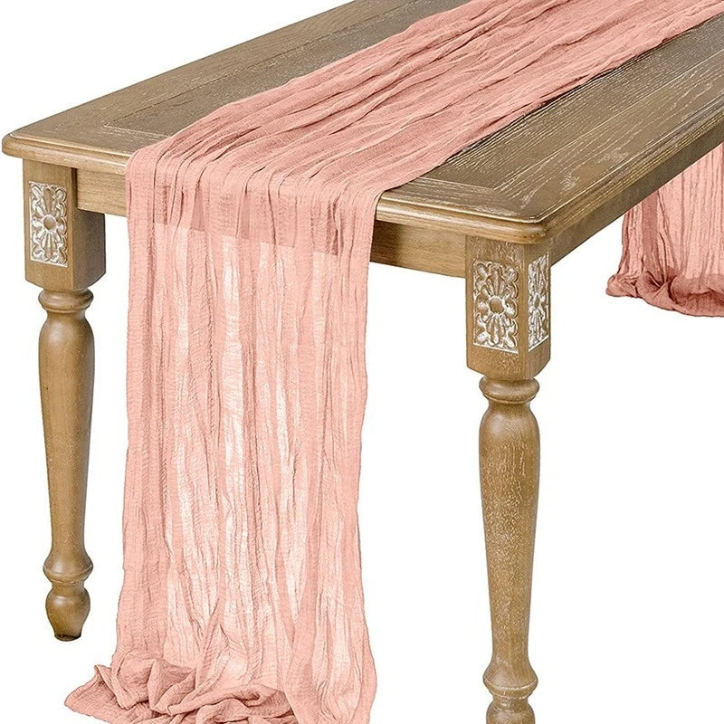 A flowing, semi-sheer gauze table runner in a dusty rose color draped over a wooden table.