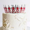 A white cake decorated with a crystal rhinestone crown topper featuring red gemstones and gold accents.