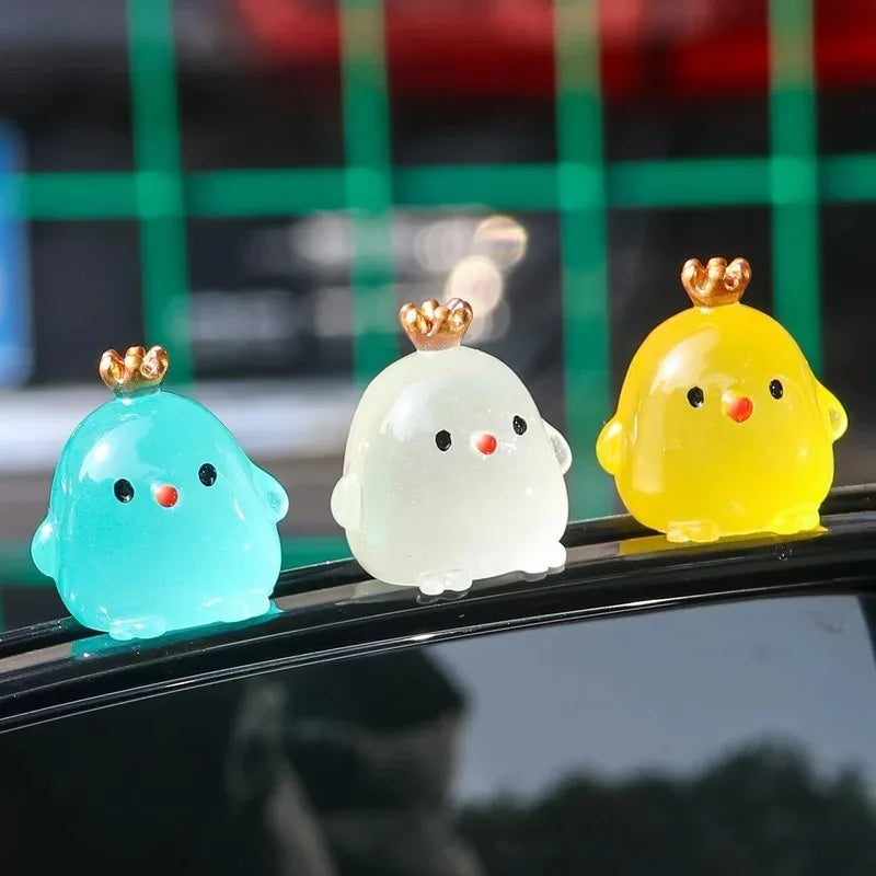 Three luminous resin chicken car ornaments in blue, white, and yellow sitting on a dark surface.