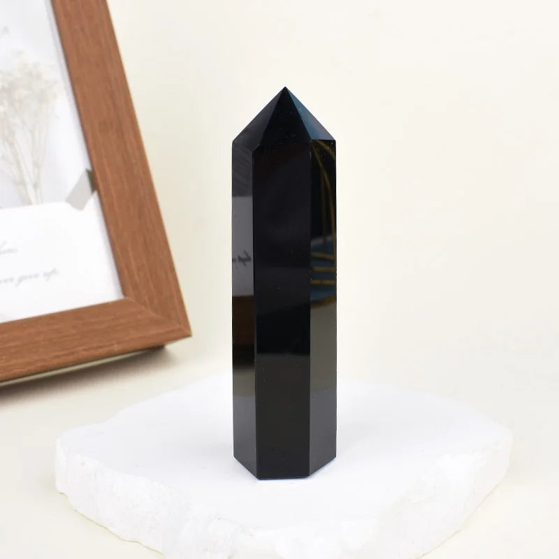 A black natural obsidian crystal obelisk point standing on a white surface next to a wooden frame.