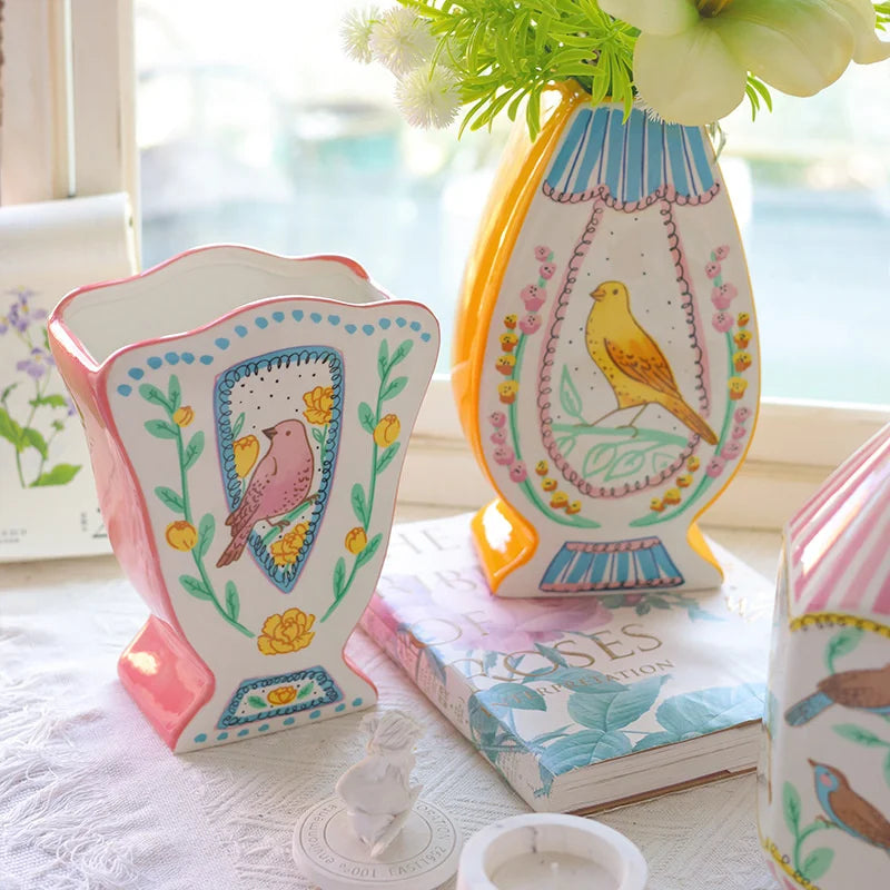 A pink ceramic bird floral vase with yellow accents sits on a white surface next to a stack of books.