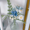 A handmade crochet flower bouquet featuring blue and white flowers in a clear glass vase on a white shelf.