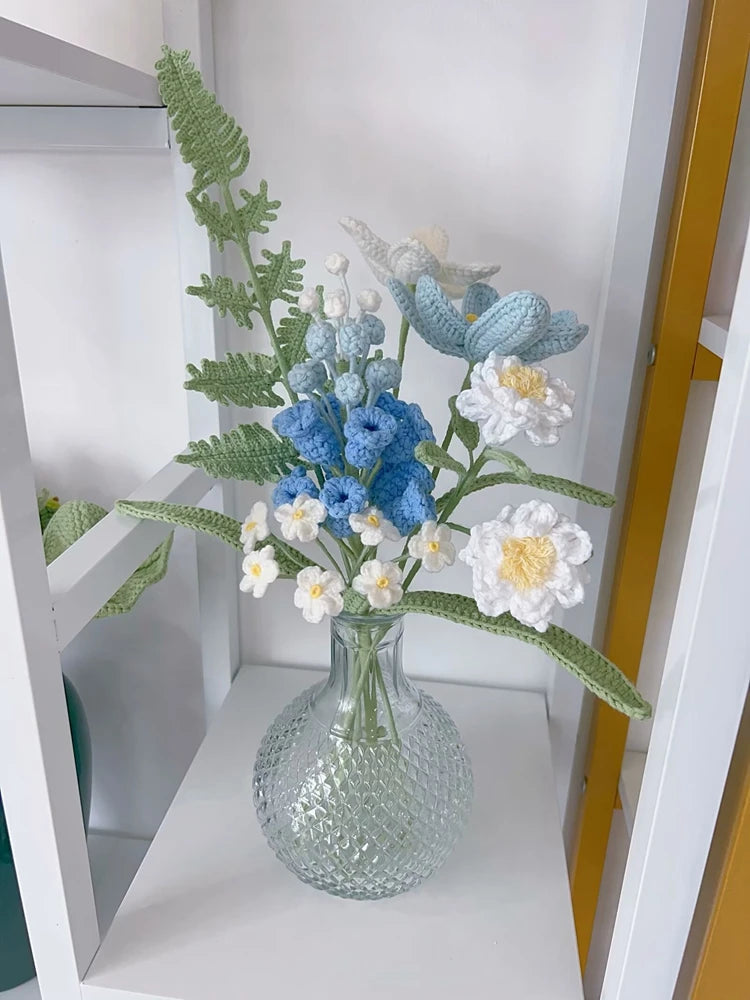 A handmade crochet flower bouquet featuring blue and white flowers in a clear glass vase on a white shelf.