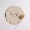 A round, light wood plaque with the cursive text hello im here engraved on it, accompanied by a dried feather.