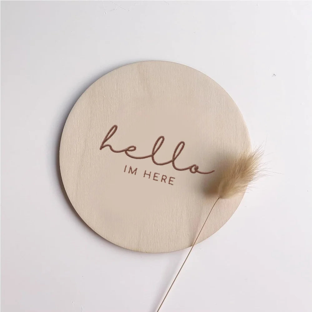 A round, light wood plaque with the cursive text hello im here engraved on it, accompanied by a dried feather.