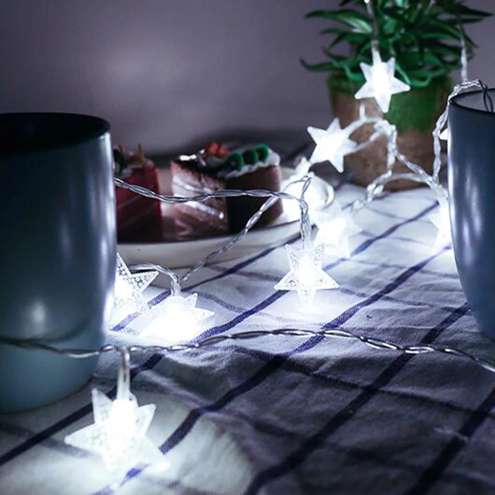 String lights shaped like stars illuminate a plaid fabric next to a dark mug and greenery.