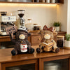 A brown cow figurine dressed as a barista, holding a cup of coffee, sitting on a wooden desk.