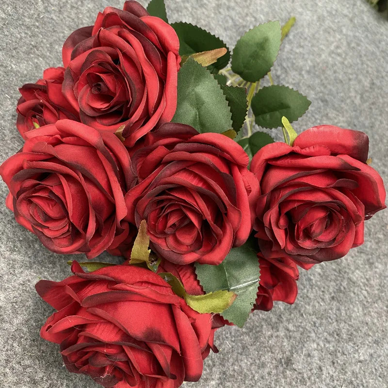 A bouquet of nine realistic red silk roses with green leaves arranged on a gray background.