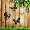 Three black metal butterfly wall art pieces hanging on a wooden plank with green vines above and grass below.
