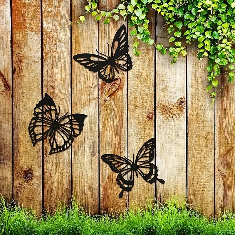 Three black metal butterfly wall art pieces hanging on a wooden plank with green vines above and grass below.