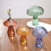 Trois vases en forme de champignons en verre, roses, jaunes et bleus, posés sur une surface en bois.