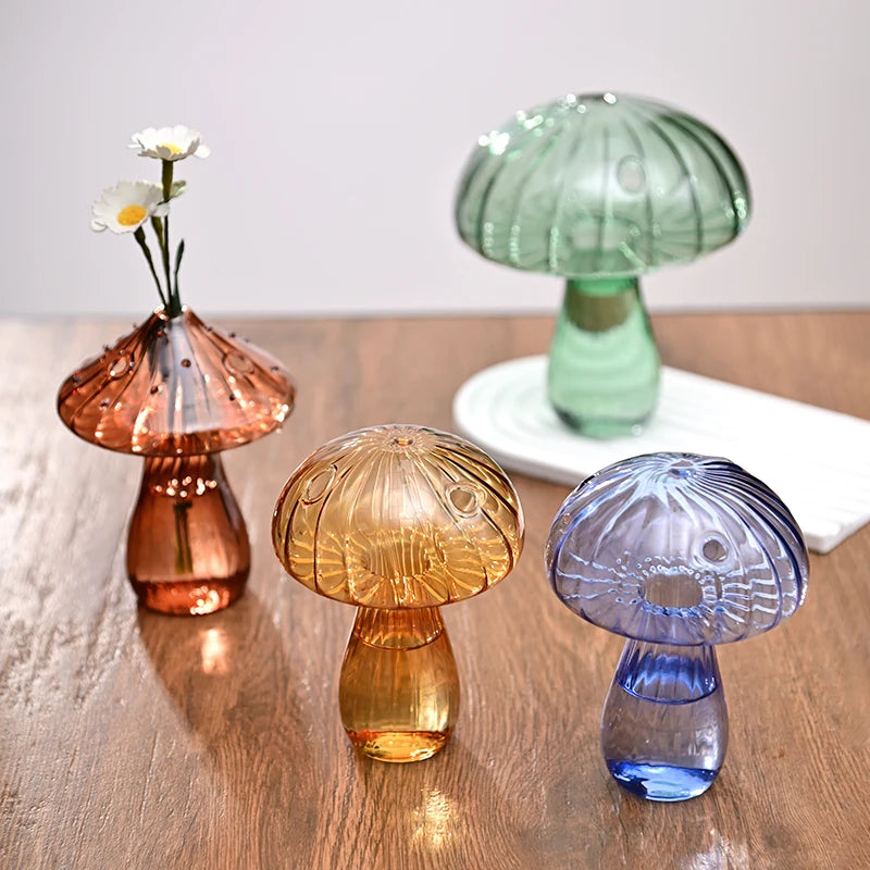 Trois vases en forme de champignons en verre, roses, jaunes et bleus, posés sur une surface en bois.