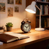 A vintage twin-bell alarm clock with a nightlight sits on a wooden desk next to a stack of books.