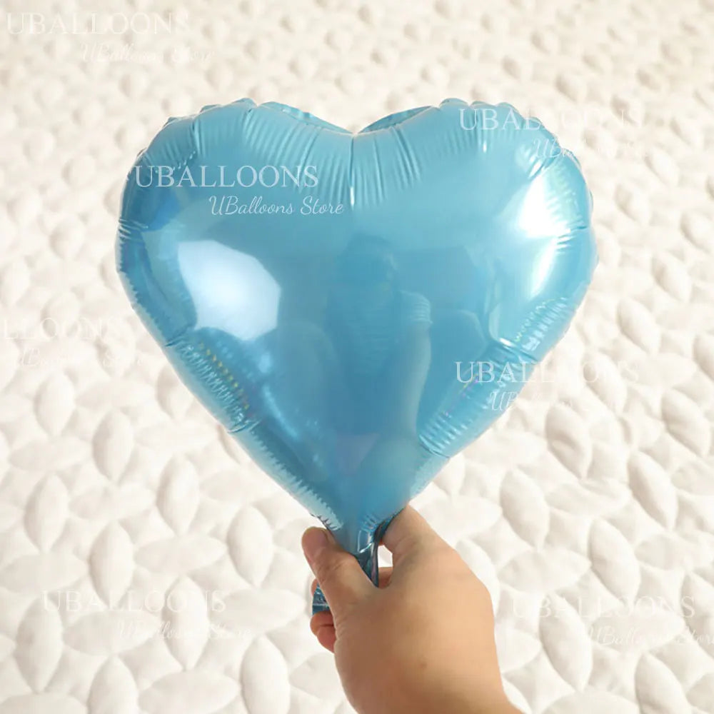A light blue heart-shaped foil balloon is being held by a hand on a white textured surface.