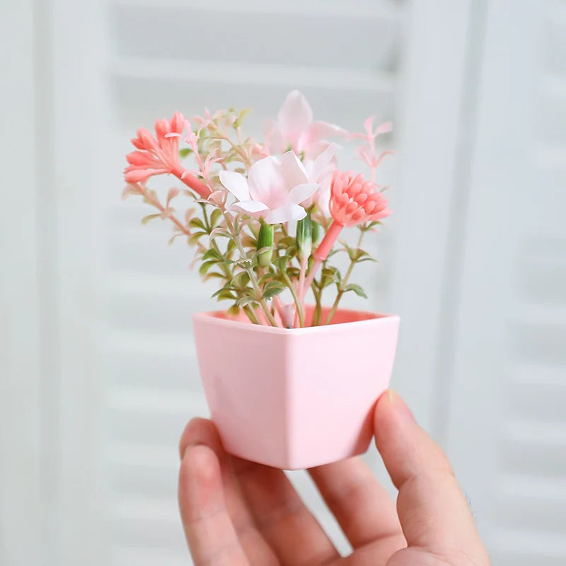 A hand holds a small pink pot containing artificial flowers with pink and coral blooms.