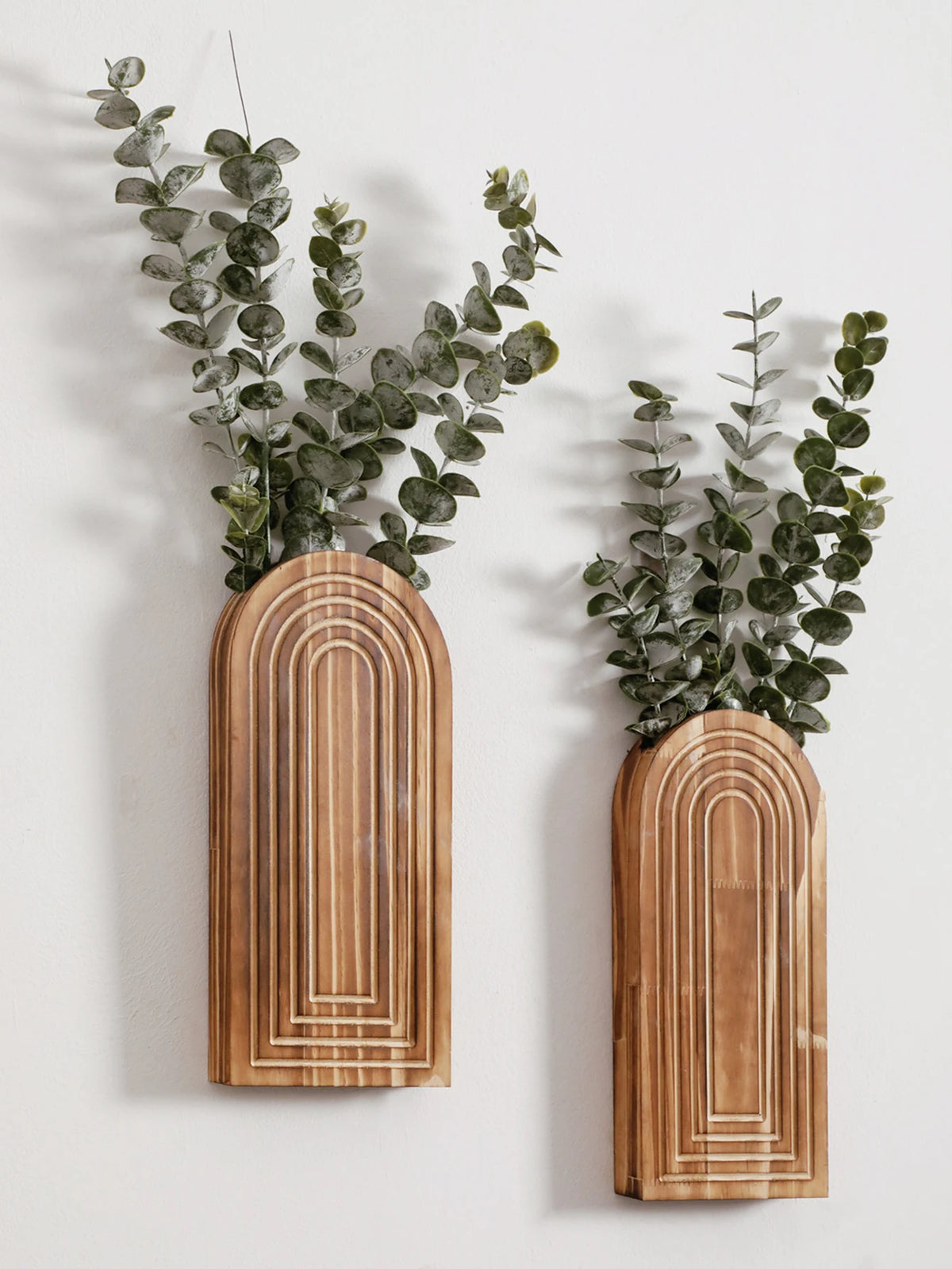 Un par de jarrones de madera arqueados con ramas de eucalipto verde, montados en una pared blanca.
