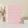 A pink laminated photo album with gold heart accents sits on a white surface next to a vase of dried flowers.