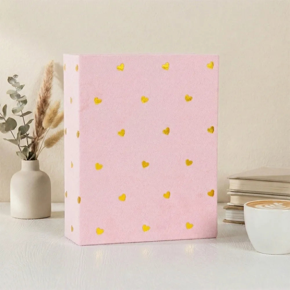 A pink laminated photo album with gold heart accents sits on a white surface next to a vase of dried flowers.
