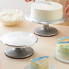 A silver aluminum cake turntable is shown with a white cake and frosting on it, next to another turntable.