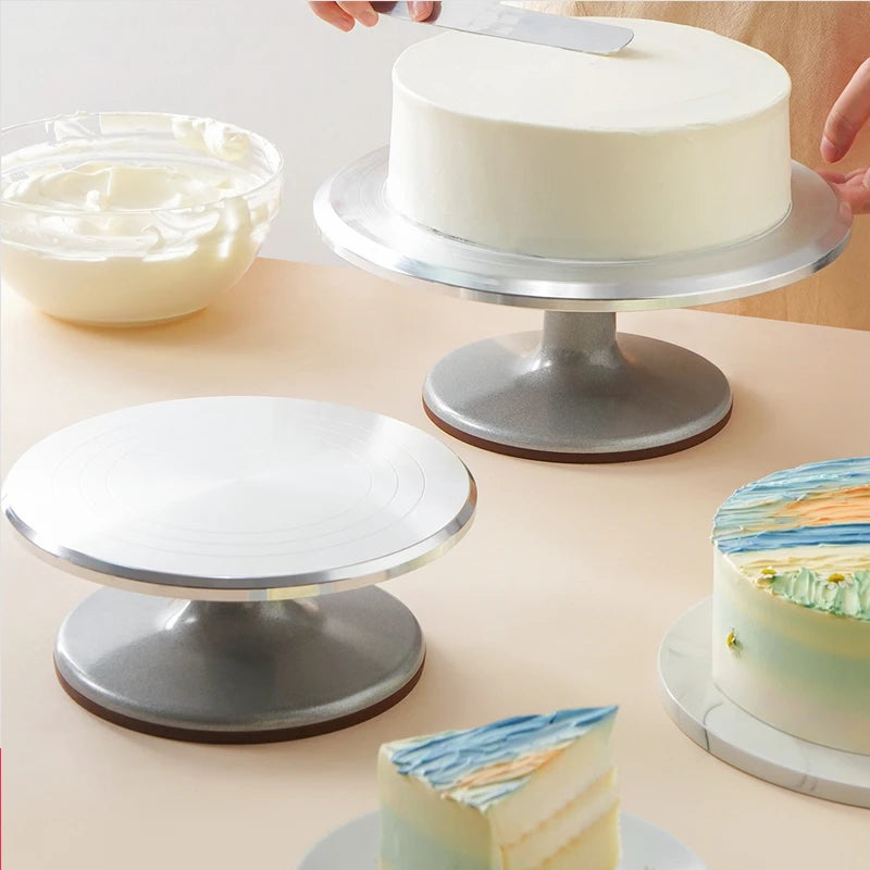 A silver aluminum cake turntable is shown with a white cake and frosting on it, next to another turntable.