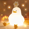 A white, glowing duck-shaped LED night light with a flower on top, resting on a wooden surface.