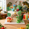 Pastel de varios niveles decorado con figuras de dinosaurios y follaje verde, con una figura de dinosaurio azul en la parte s