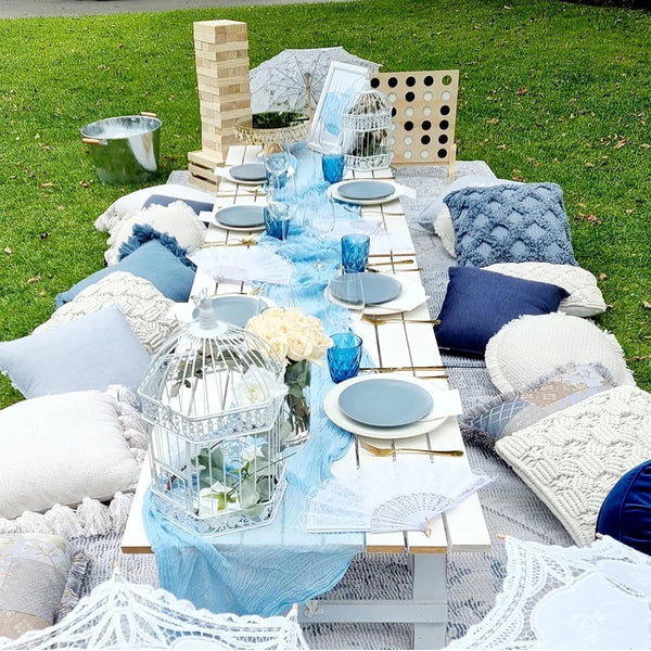 A rustic and boho table setting featuring a light blue cheesecloth runner, glassware, and white textured pillows on a green l