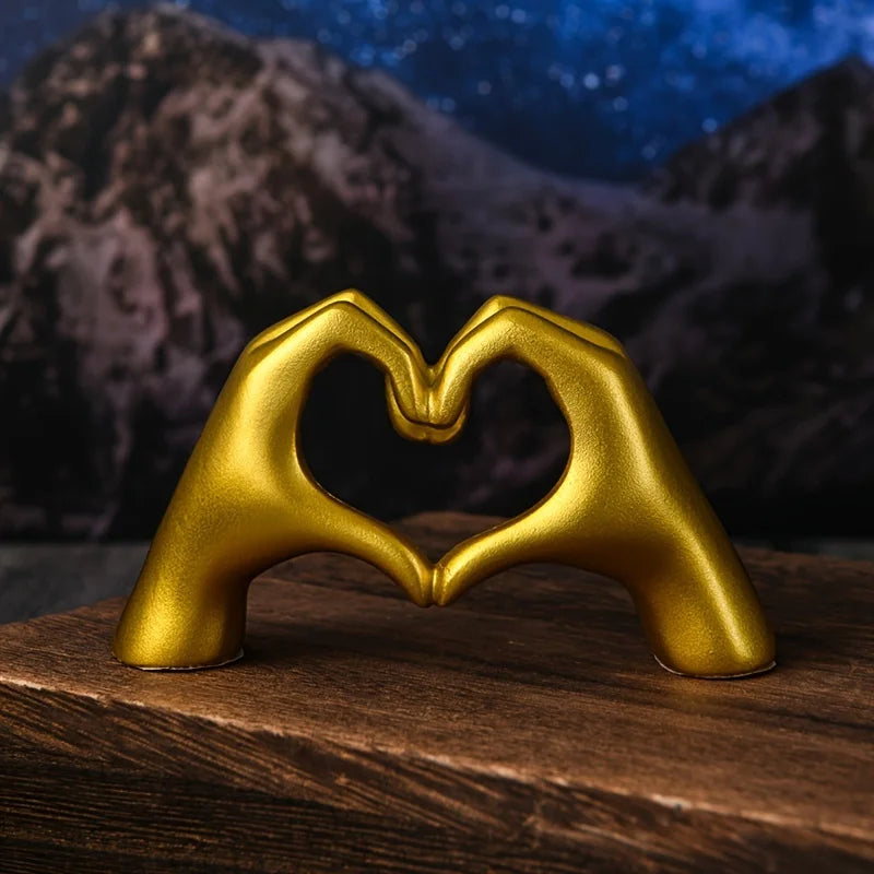 A golden heart shape sculpture made from a hand gesture, resting on a wooden surface against a dark mountain background.
