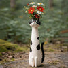 A white resin cat figurine with black spots holds a bouquet of colorful wildflowers.