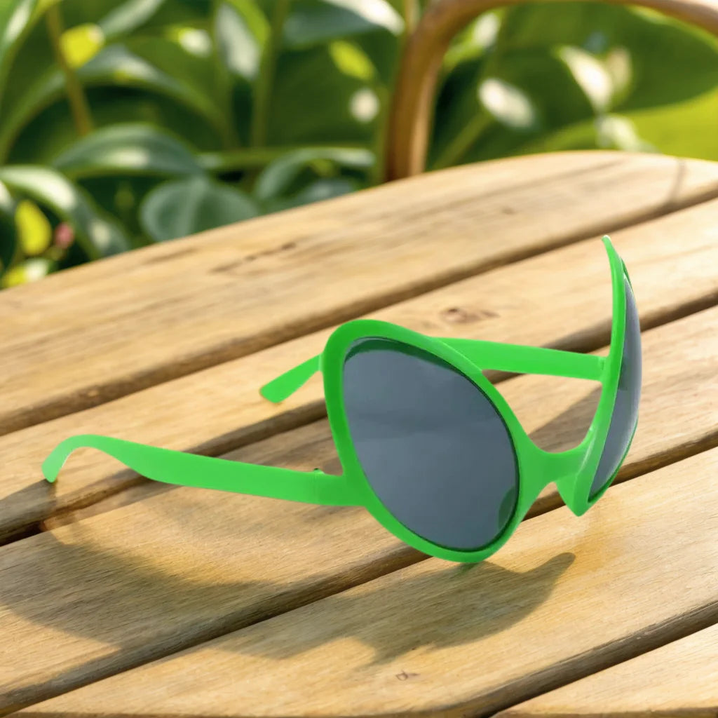 Green alien sunglasses resting on a wooden surface with blurred green foliage in the background.