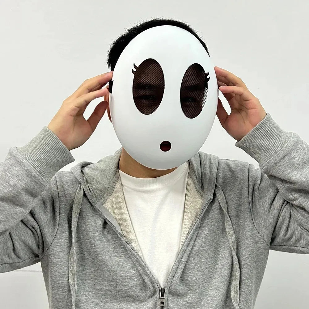 A person is holding a white, round mask resembling a shy guy character with large black eye holes and a small red nose.