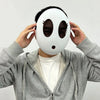A person is holding a white, round mask resembling a shy guy character with large black eye holes and a small red nose.