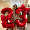A woman holds large red foil number balloons shaped like the number 32 with matching bows.