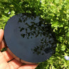 A hand holds a smooth, dark obsidian mirror reflecting green foliage in natural sunlight.