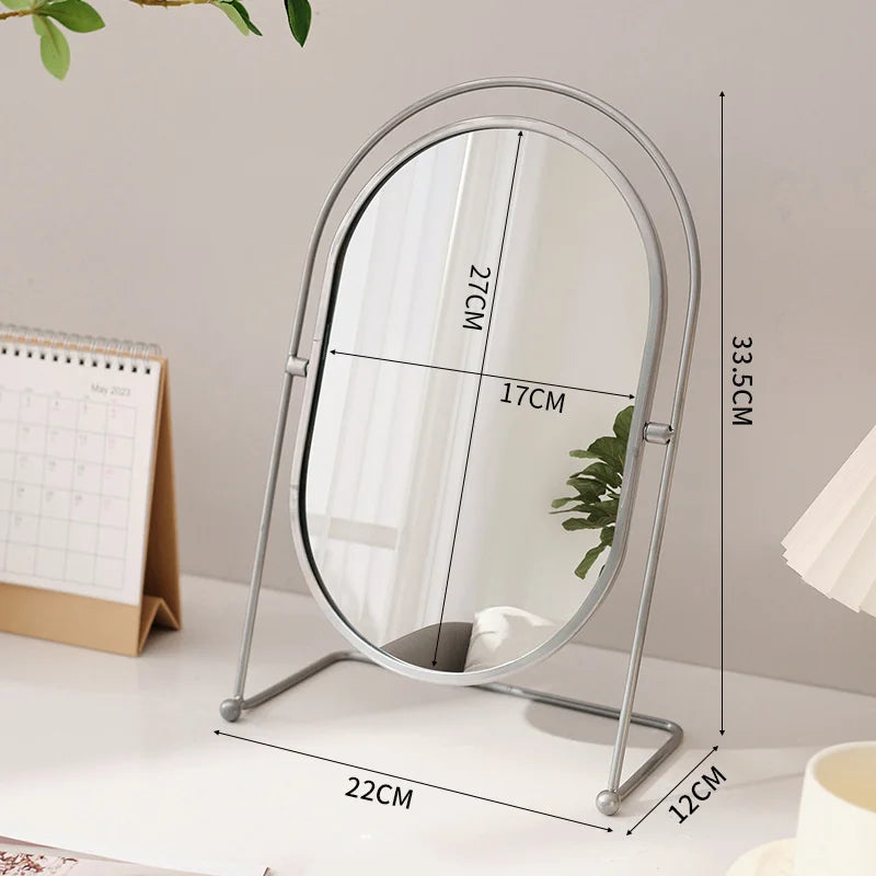 A silver oval vanity mirror with a rotating frame and a stand is shown on a white surface.