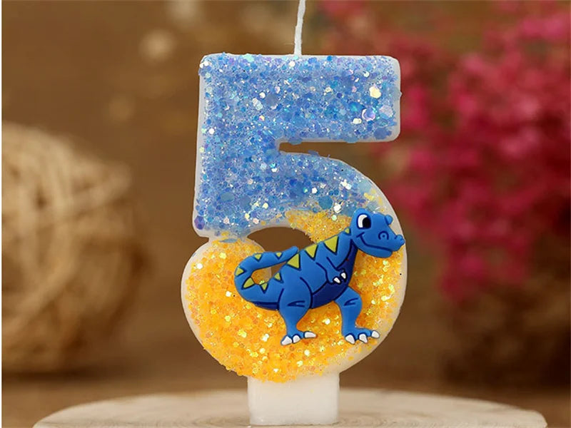 A birthday candle shaped like the number 5, featuring blue glitter and a small blue dinosaur on an orange base.