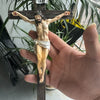 A resin sculpture of the crucified Christ on a wooden cross, held by a hand against a green plant background.
