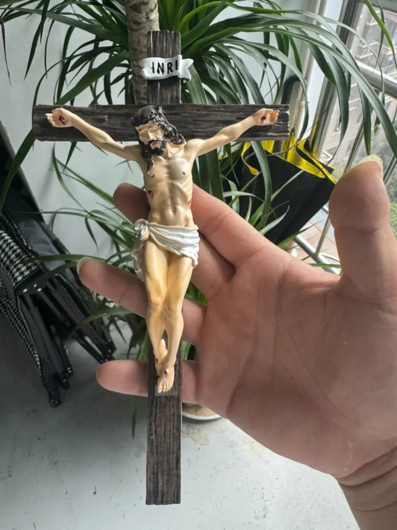 A resin sculpture of the crucified Christ on a wooden cross, held by a hand against a green plant background.