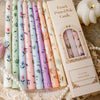 A set of French printed long stick candles in various pastel colors displayed on a lace cloth.