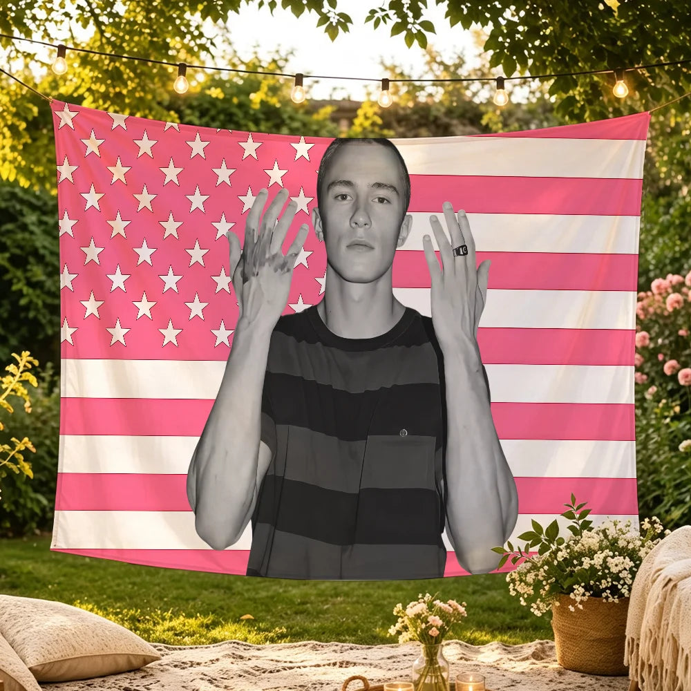 A young man stands in front of a pink and white American flag tapestry outdoors.