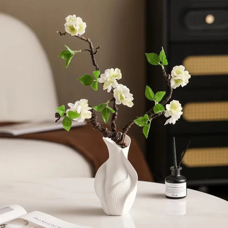 A white ceramic vase with an abstract grass motif holds a branch of flowering greenery on a white table.