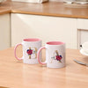 A white ceramic coffee mug with a pink handle featuring a gold letter initial decorated with red and green flowers.