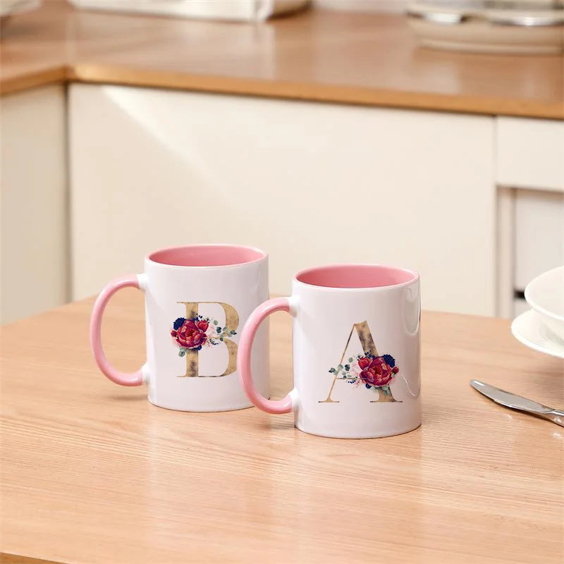 A white ceramic coffee mug with a pink handle featuring a gold letter initial decorated with red and green flowers.