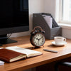 A vintage twin-bell alarm clock with a wooden casing sits on a desk next to a cup of coffee and a notebook.