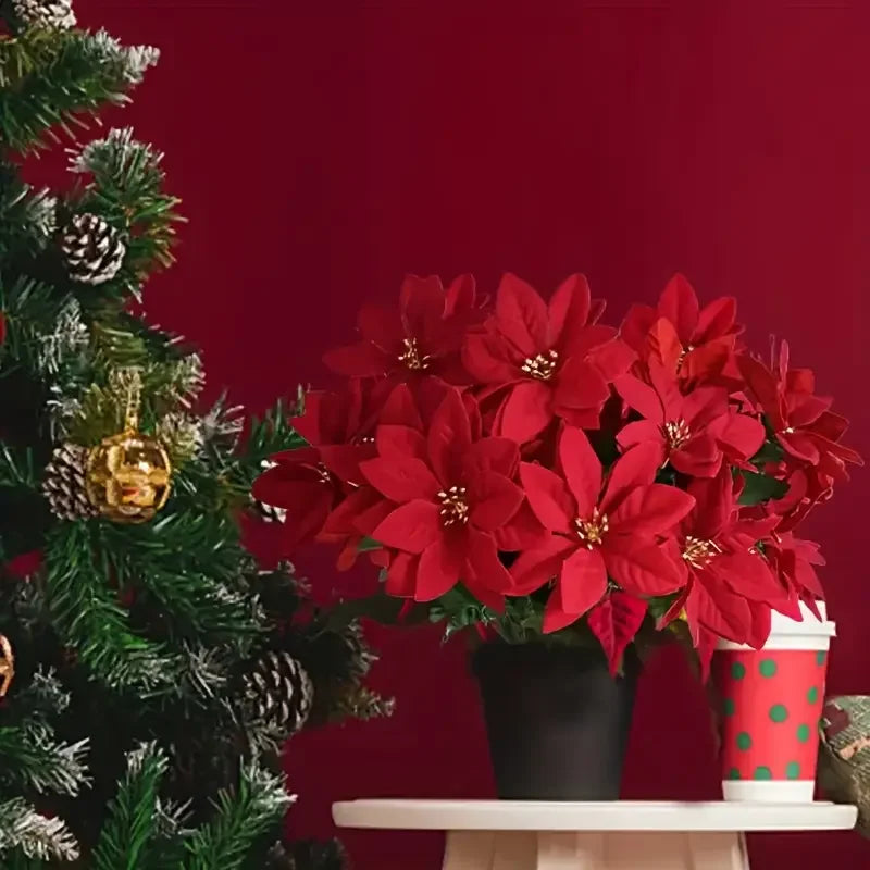 A bouquet of realistic artificial red poinsettias in a black pot displayed on a white surface next to a Christmas tree.